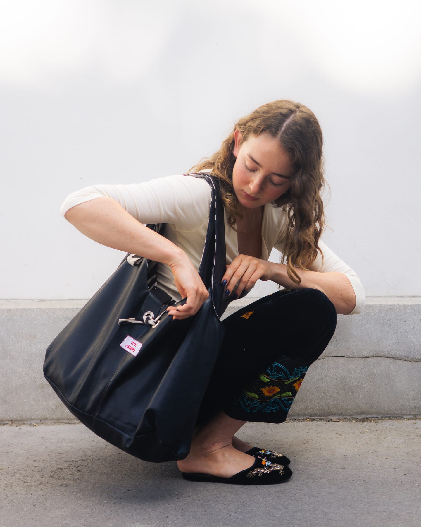 Beth Studio: model crouching and reaching into a large black tote bag, in embroidered trousers and black slippers.