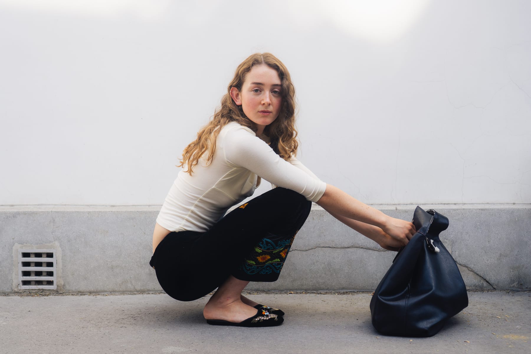 Model in a cream top and black trousers crouching beside a black tote bag against a white wall, Beth Studio.