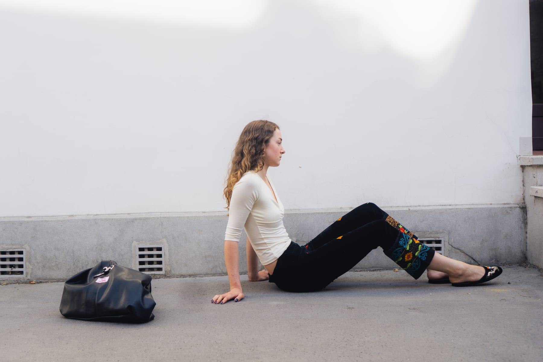 Model sitting on the pavement against a white wall, a black Beth Studio bag beside her.