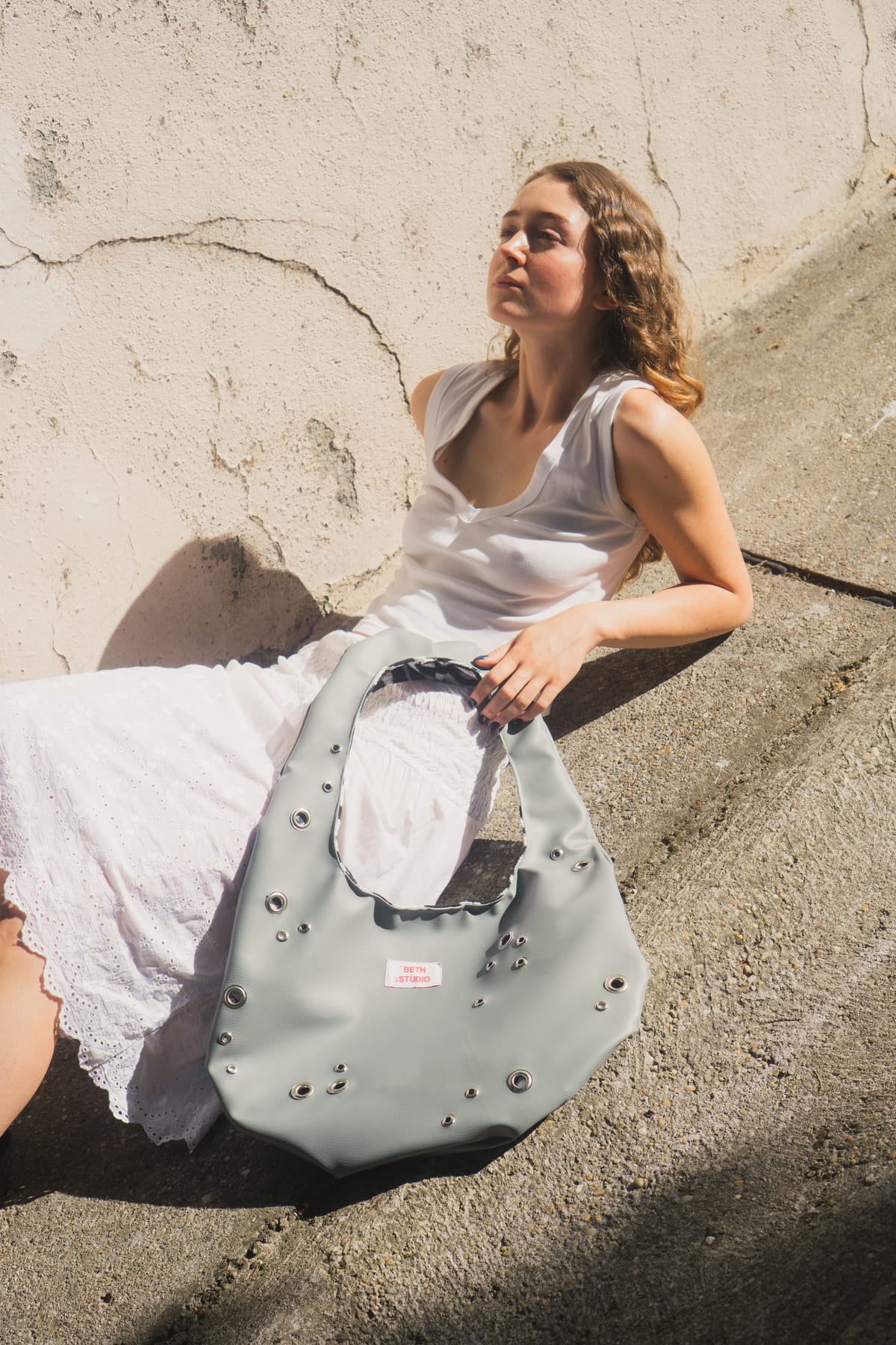 Model in a white dress sitting on a ledge in the sun, holding a grey studded Beth Studio bag.