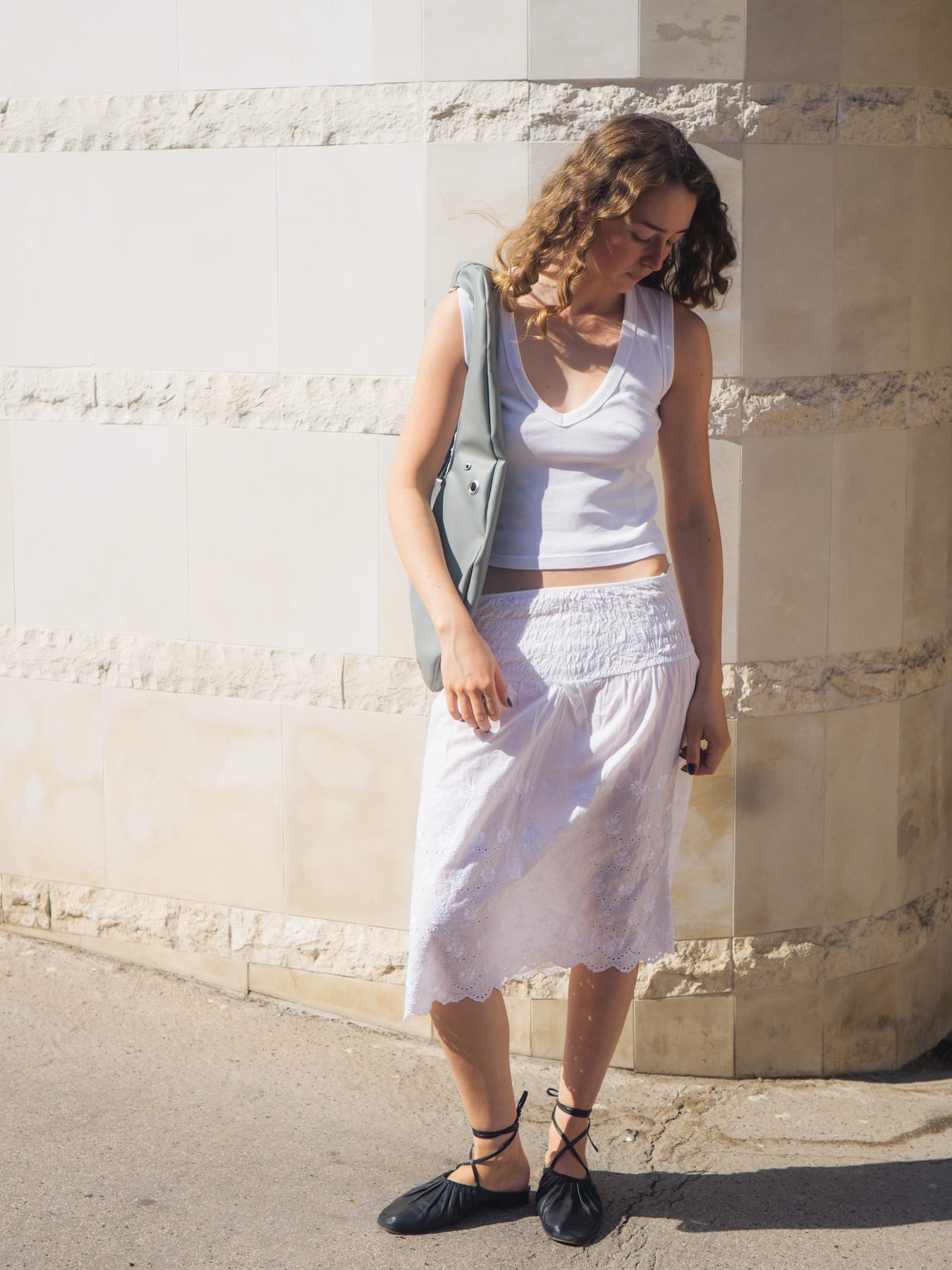 Model in a white top and skirt by a pale stone wall, a grey bag on her shoulder, looking down.