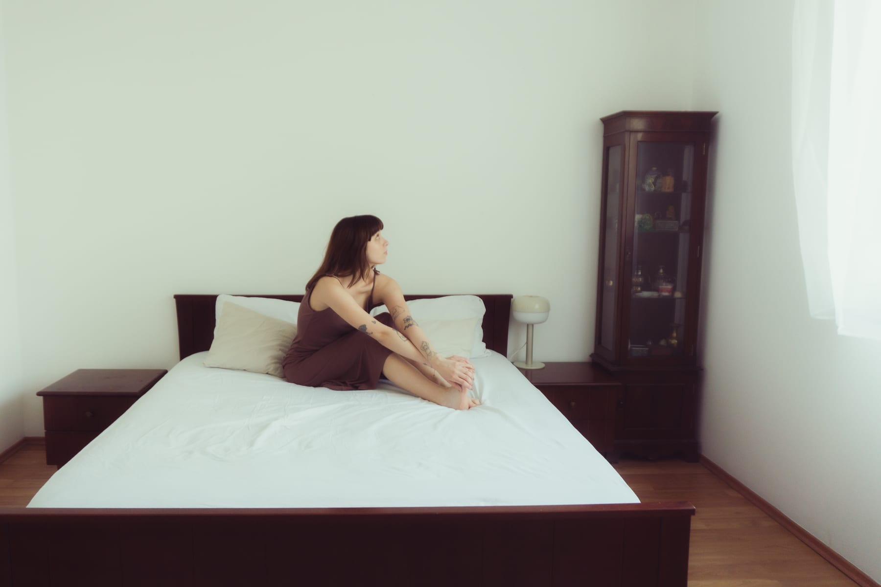 Bogi sitting cross-legged on a white double bed in a bright bedroom with a dark wooden cabinet.