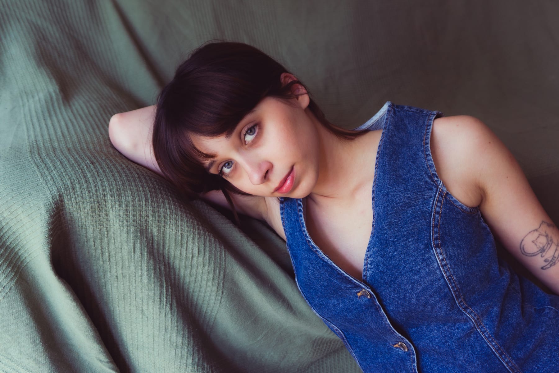 Bogi in a denim waistcoat lying on a green blanket, one hand behind her head, looking up.