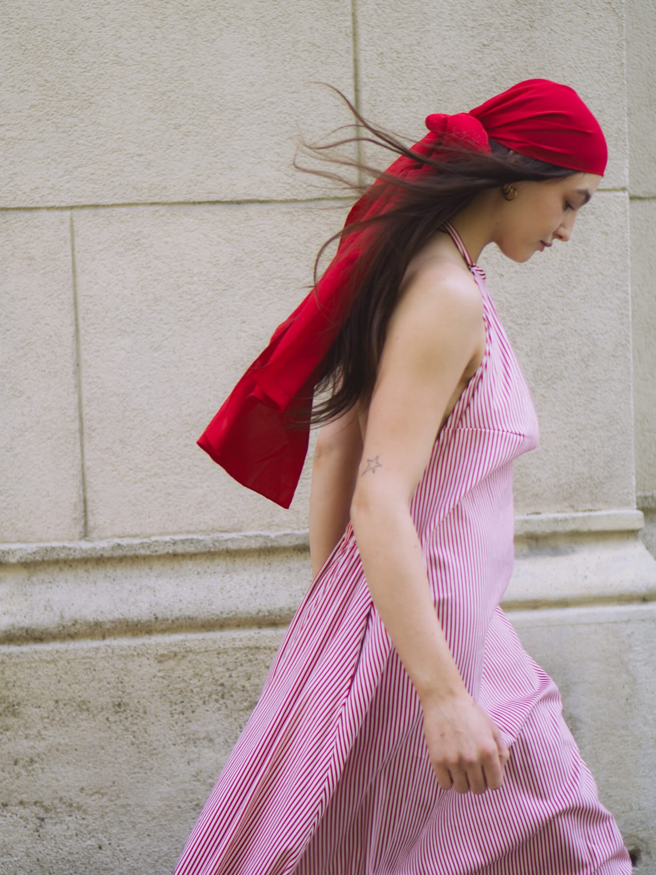 Celestial by Karácsony Andrea: model in a pink striped dress and a red headscarf walking past a stone wall.