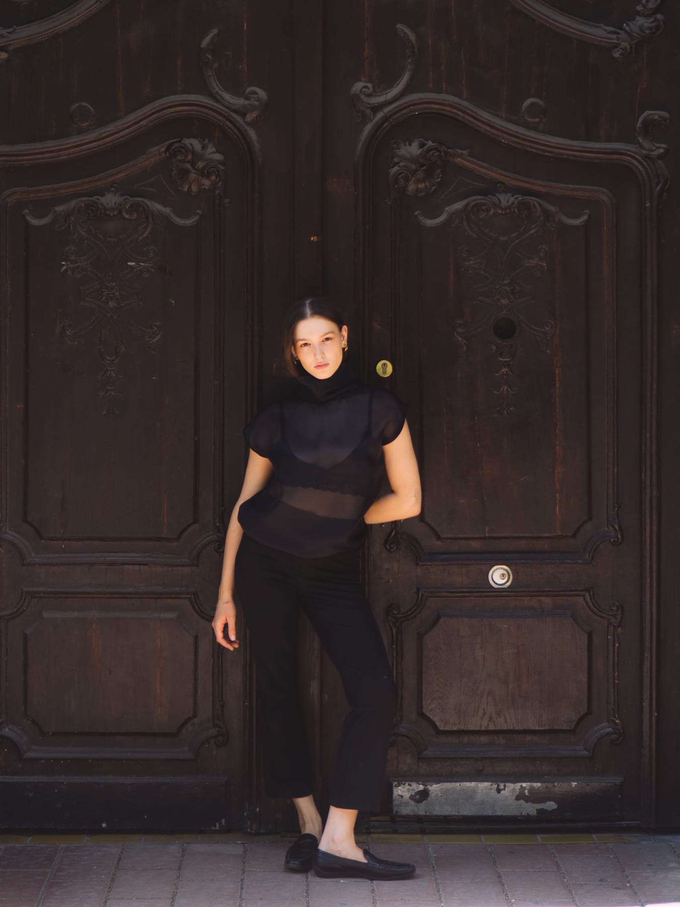 Model in a black sheer top and trousers standing before a carved dark wooden gate, Celestial by Karácsony Andrea.