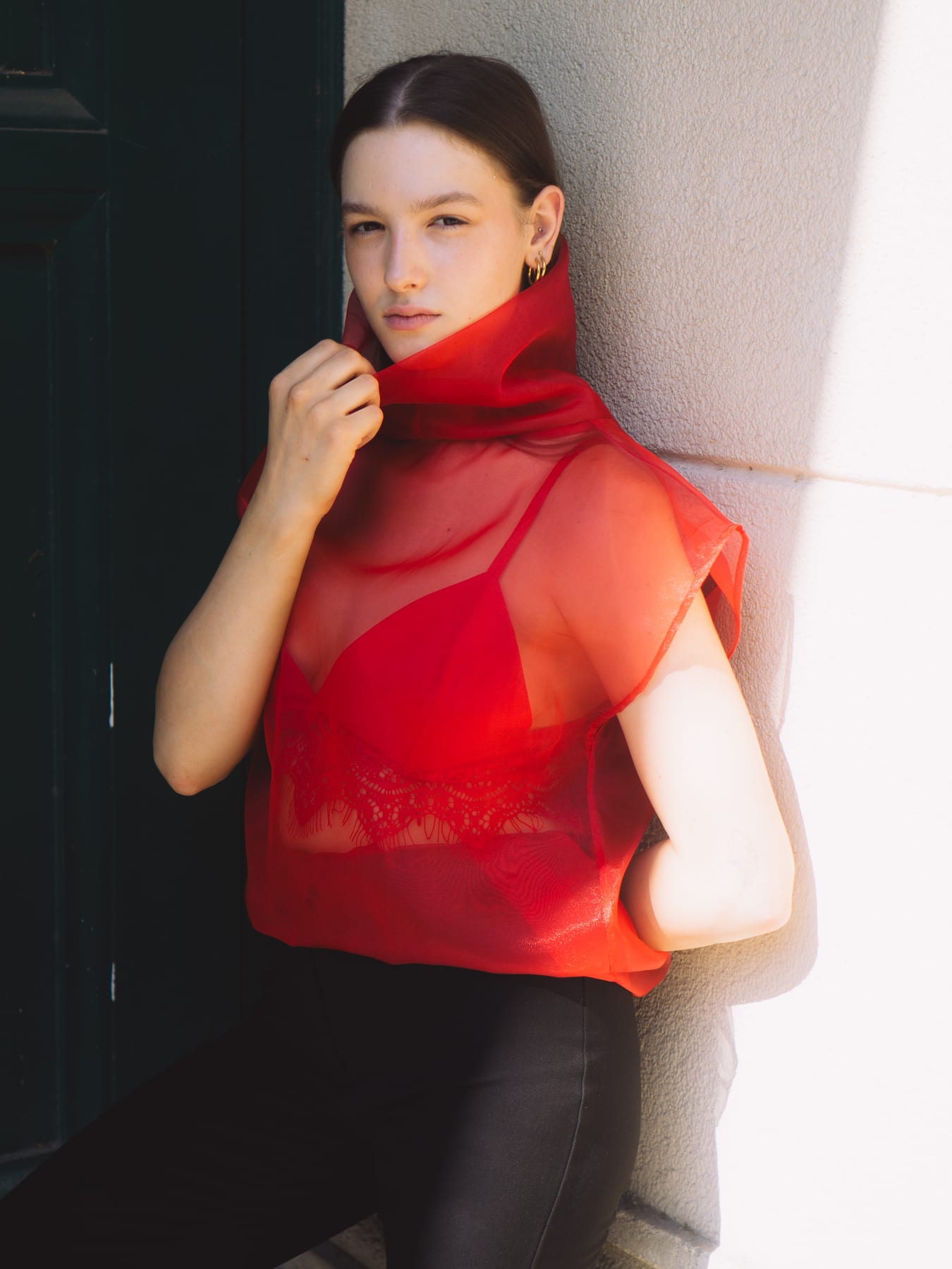 Model in a sheer red organza top with a high collar leaning against a sunlit pale wall.