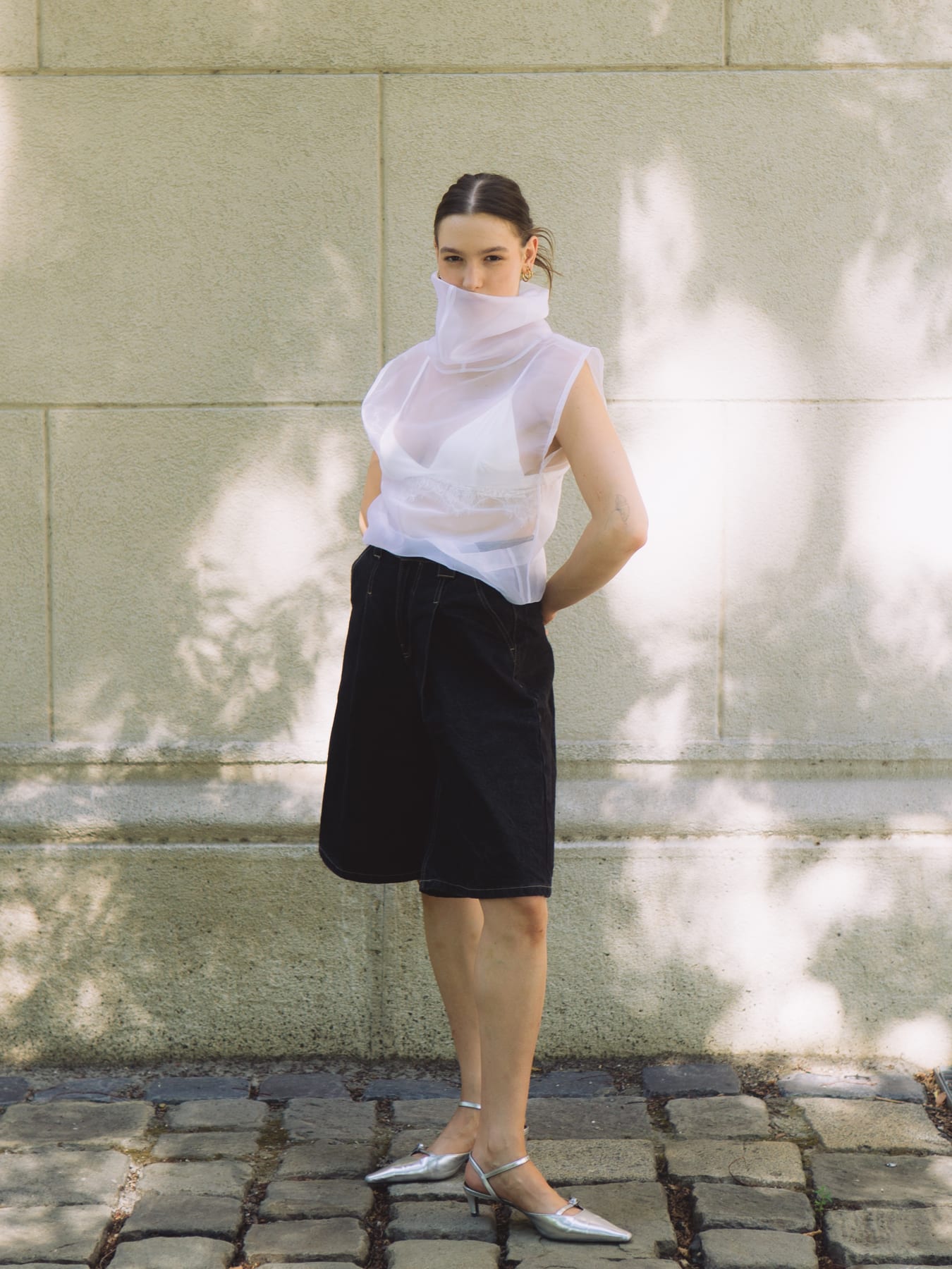 Model in a sheer white high-neck top and black shorts against a sunlit stone wall, silver flats, Celestial.