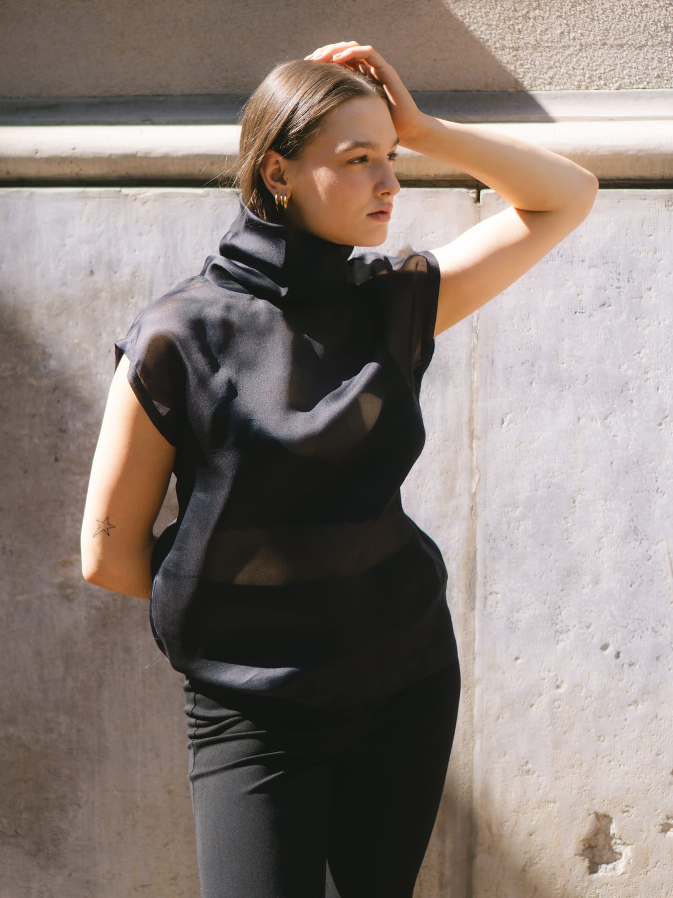 Model in a black high-neck top, one hand on her head, against a pale wall in hard sunlight.