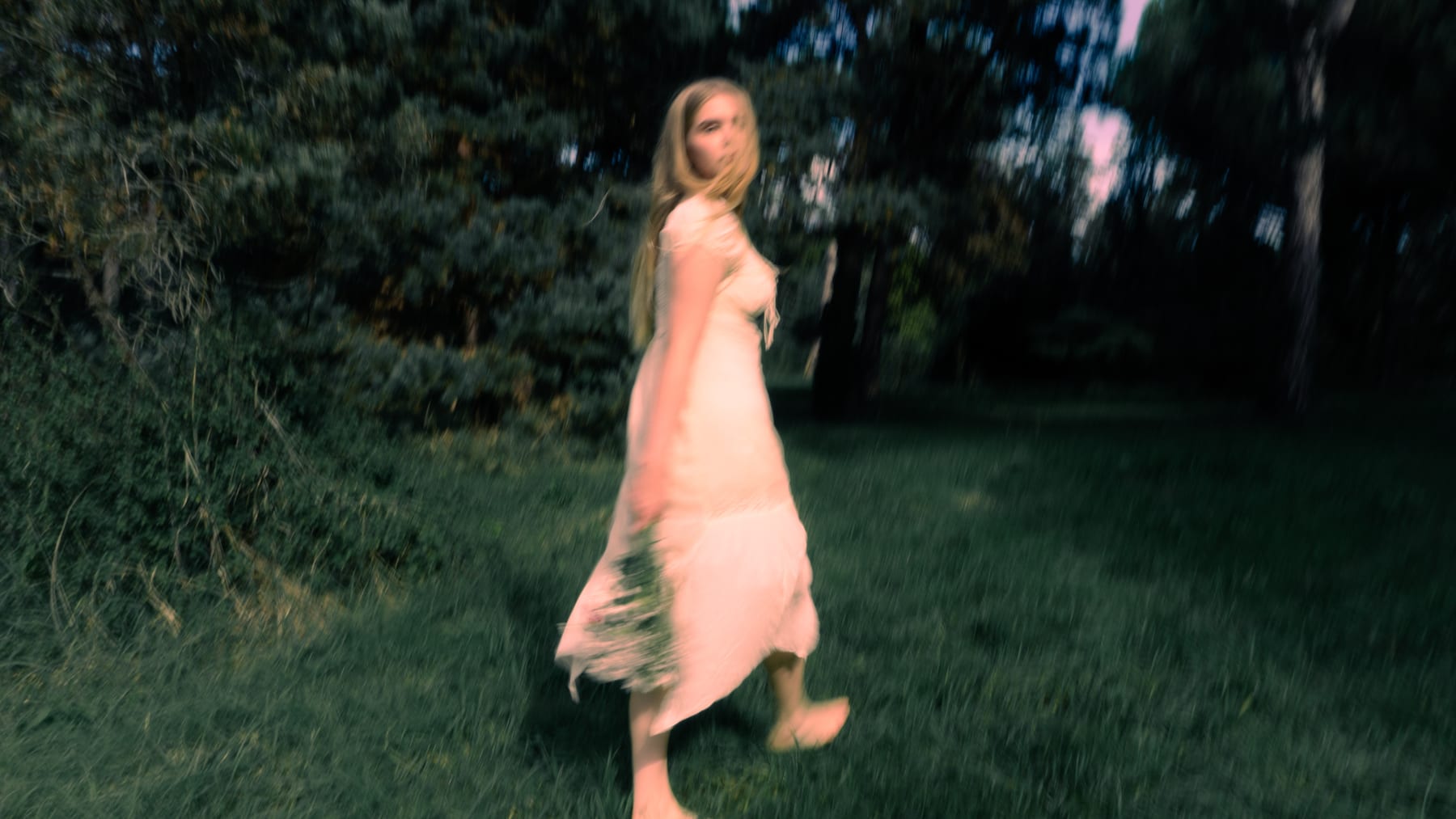 Woman in a pale dress walking through a meadow, looking back over her shoulder.