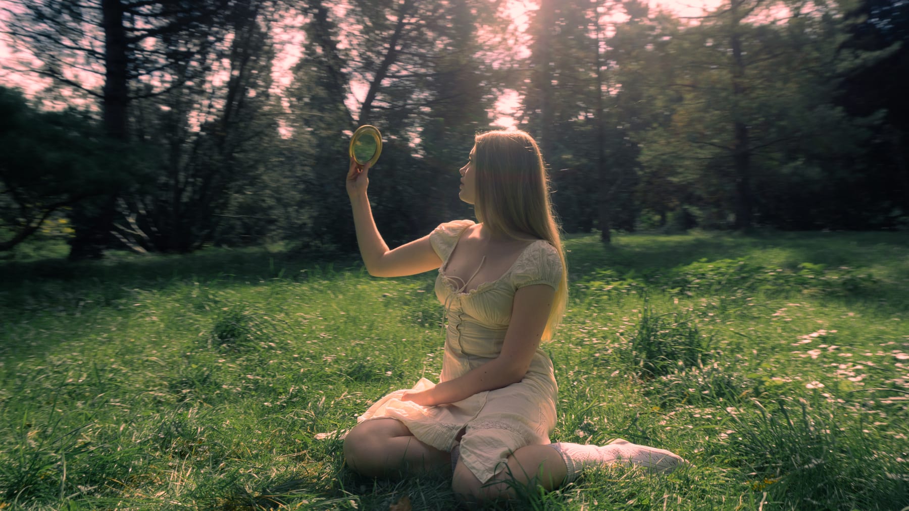 Woman with long blonde hair sitting in a sunlit meadow, holding up a small round mirror.