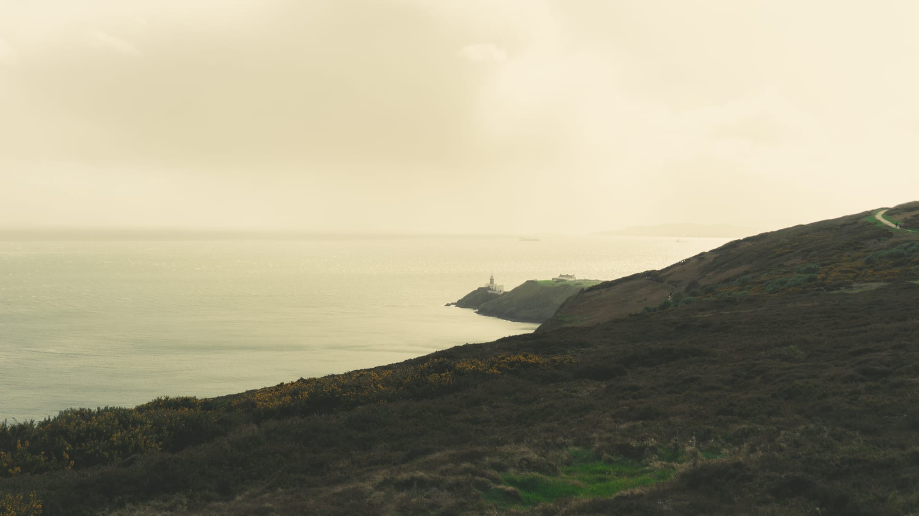 A dark, steep coastline above a hazy sea with a lighthouse in the distance, Ireland.
