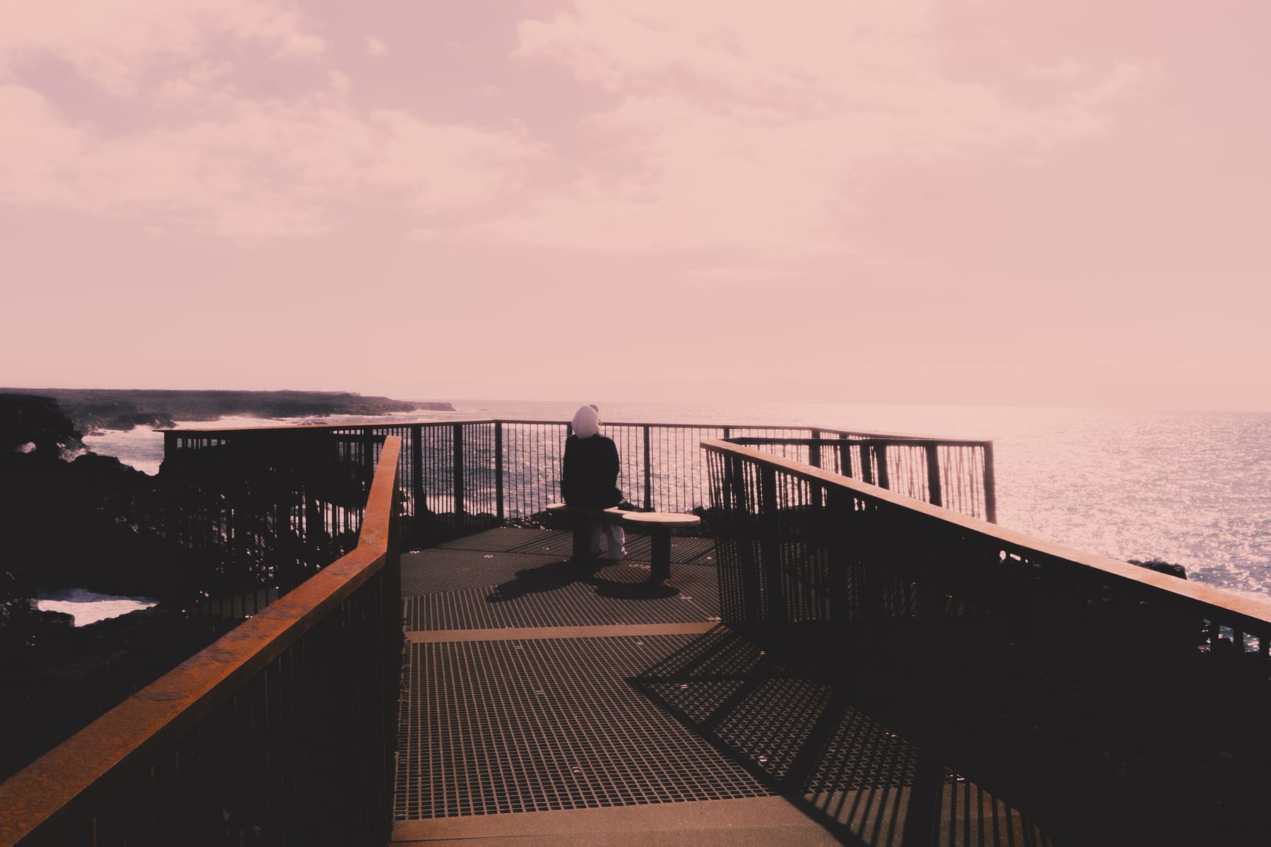 A person on a railed viewing platform above the rocky Icelandic coast, in pink evening light.