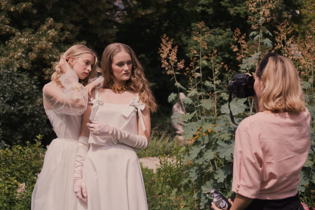 Behind the scenes: two models in white dresses in a garden, a photographer in pink filming them.