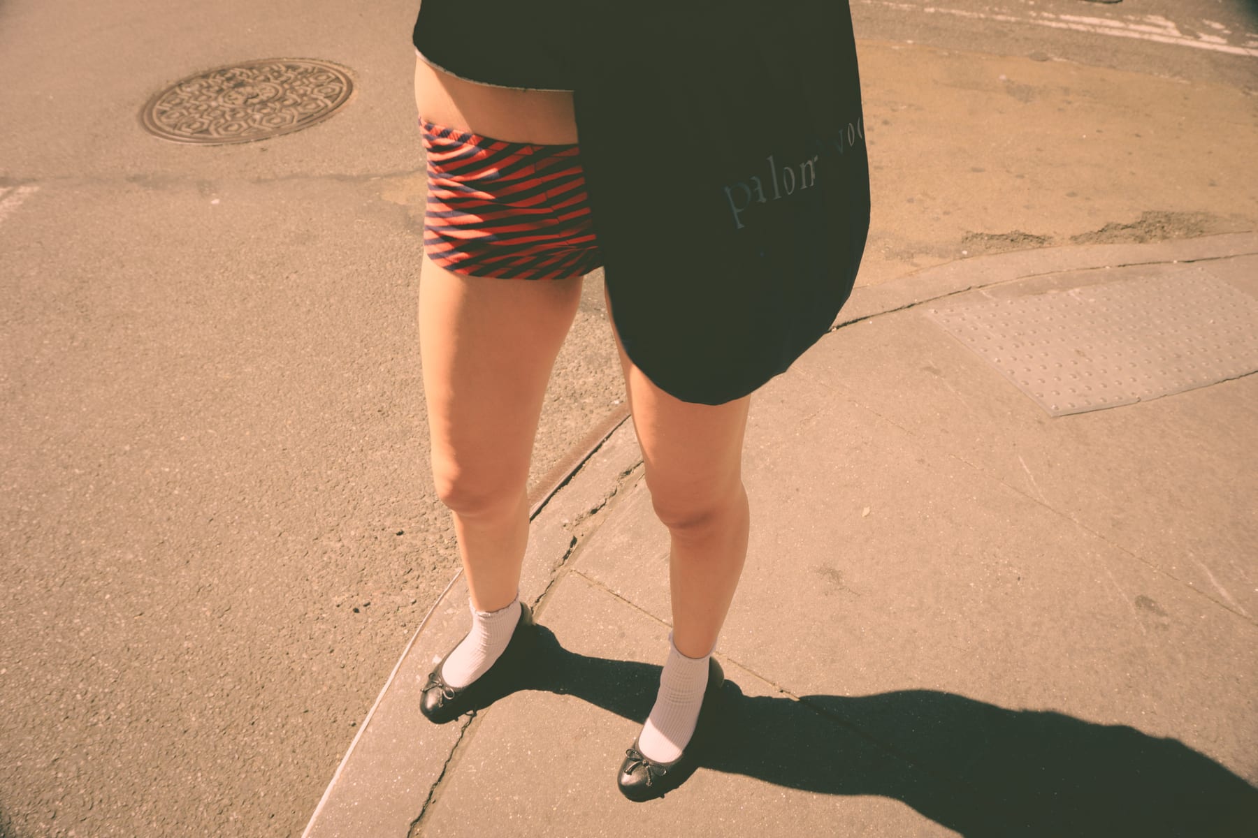 Legs in white socks and black shoes on a sunlit New York pavement, a striped skirt and a long shadow.