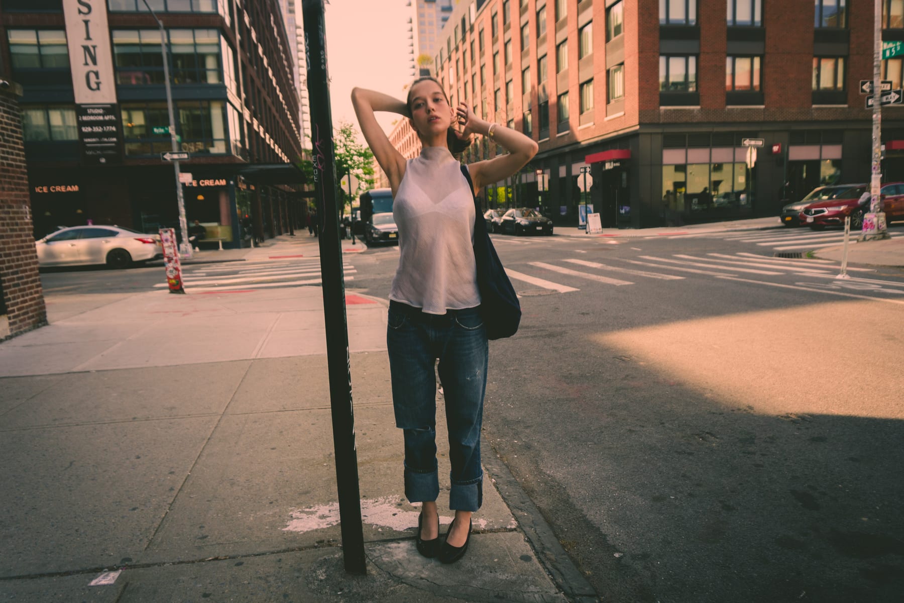 Woman in a white top and jeans by a pole on a New York street corner, hands on her head.