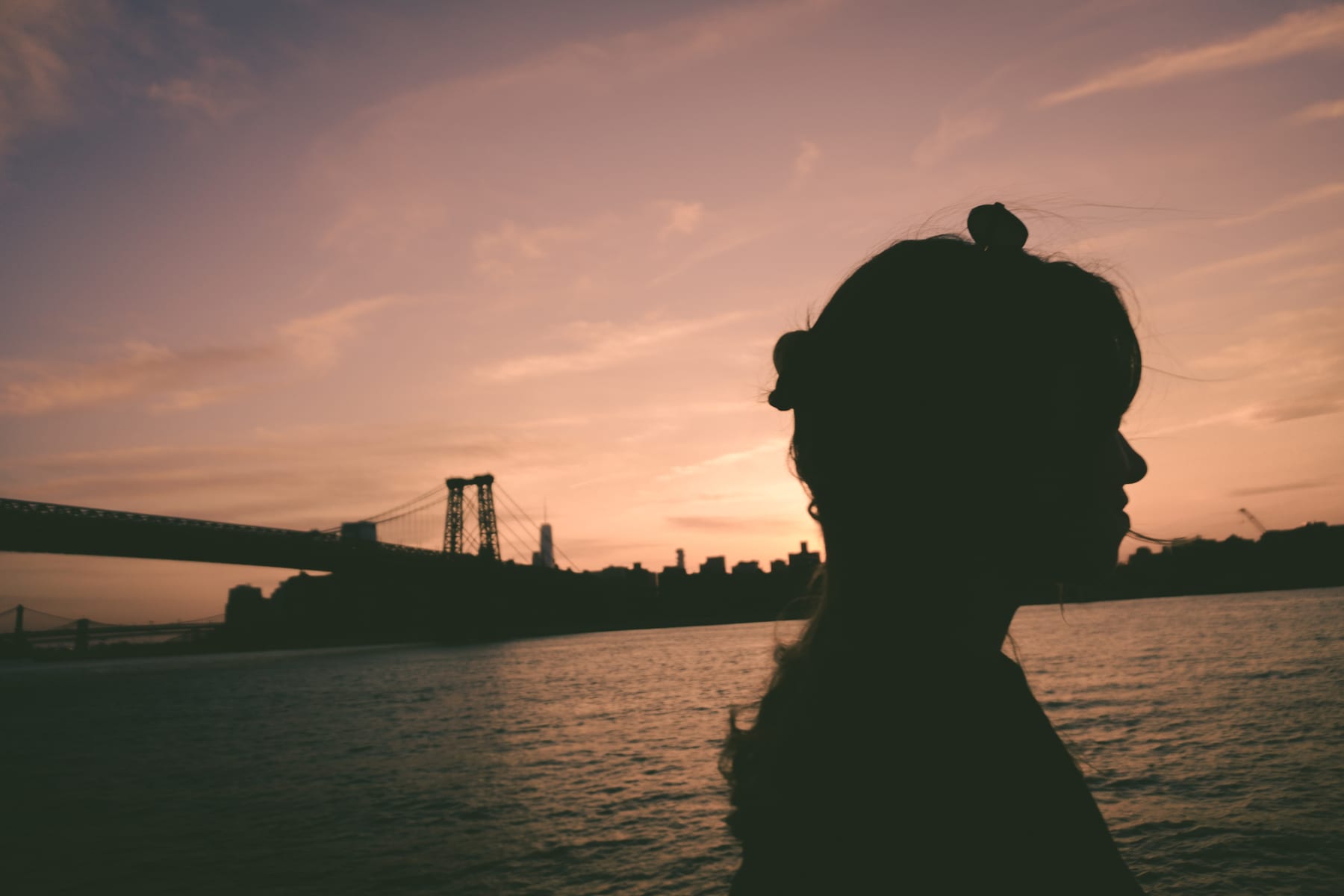 Silhouette in profile at sunset with the Williamsburg Bridge and the East River behind.