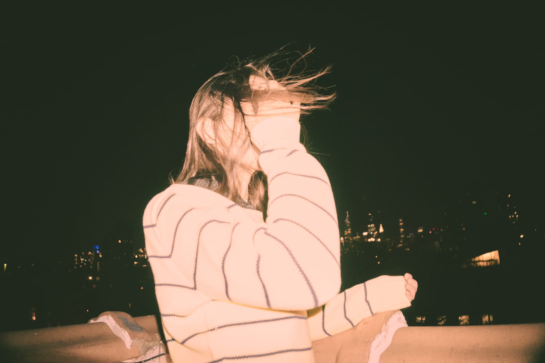 Woman in a striped sweater at night, hair blown across her face, New York city lights behind.