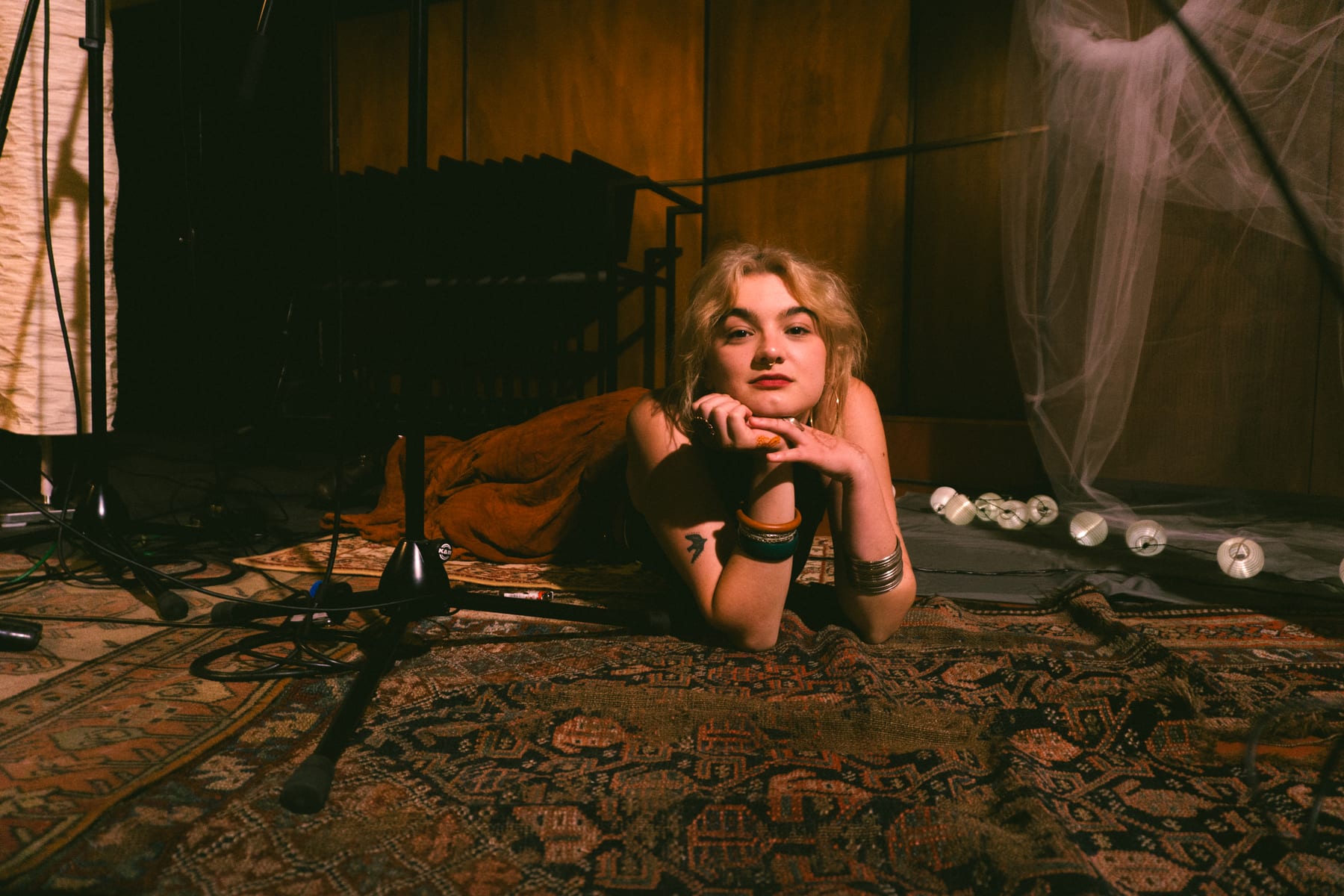 Singer lying on a patterned rug among microphone stands in a dim recording studio.