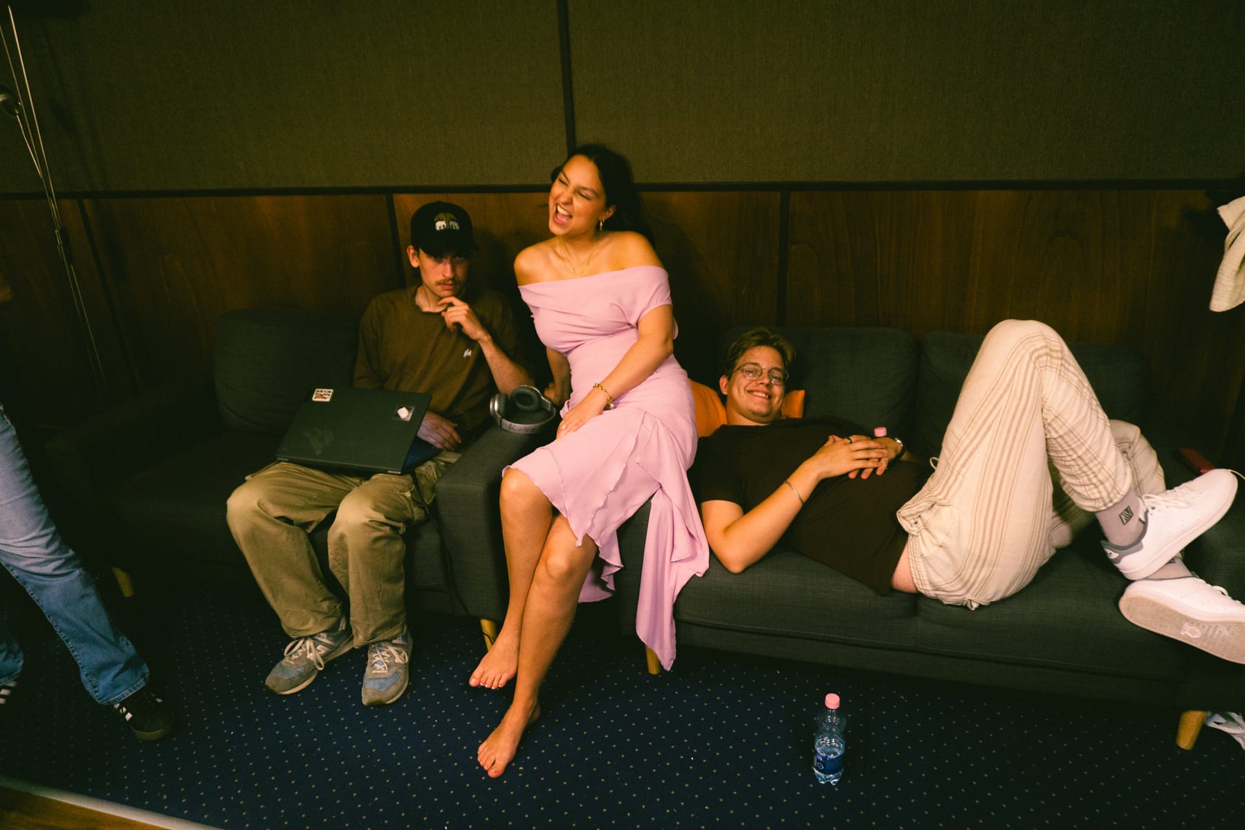 Pati Piko in a pink dress laughing with two musicians on a dark blue sofa in the studio.