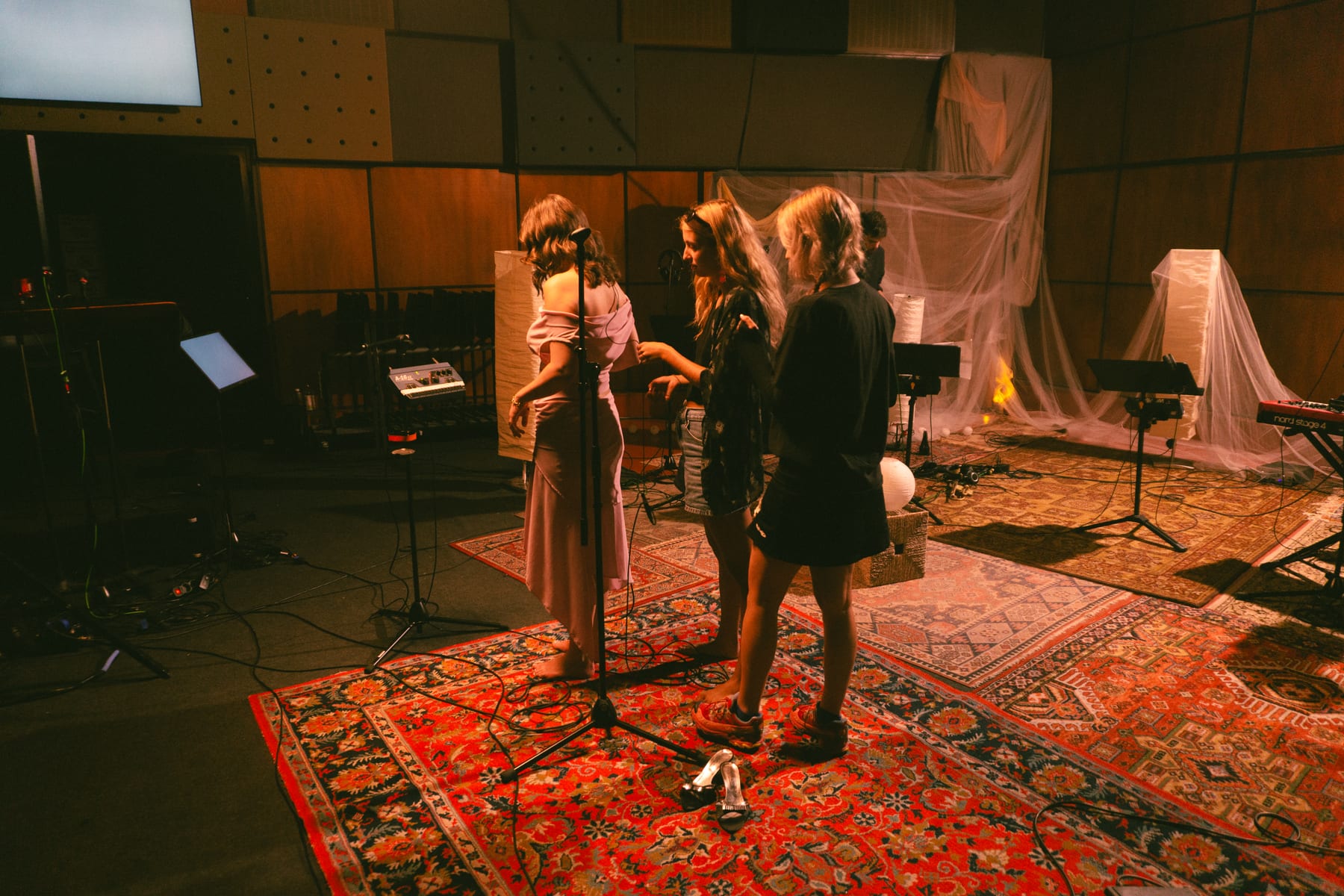 Singers and musicians rehearsing on red rugs in a warmly lit recording studio.