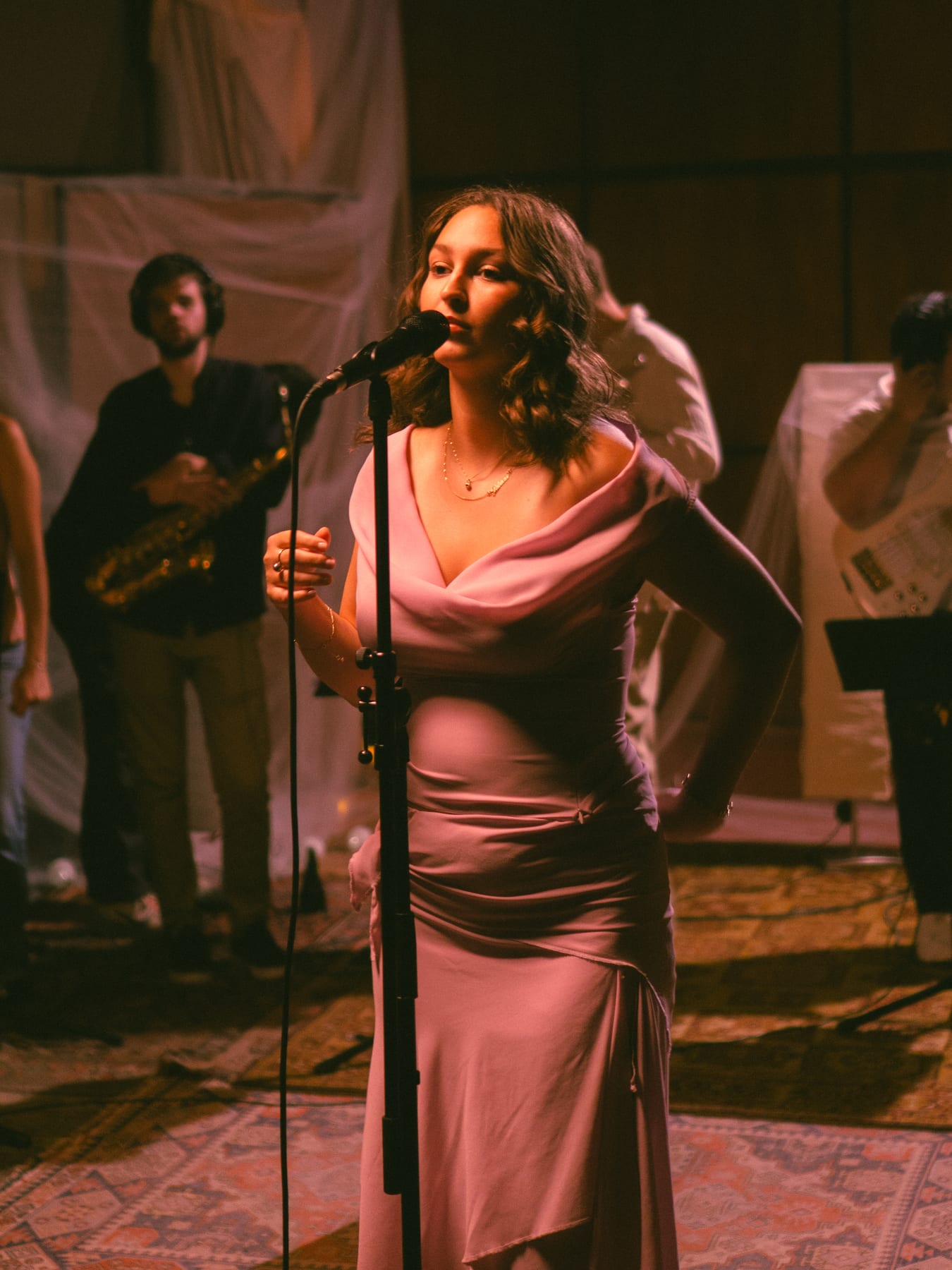 Pati Piko singing into a microphone in a pink satin dress, a guitarist behind her.