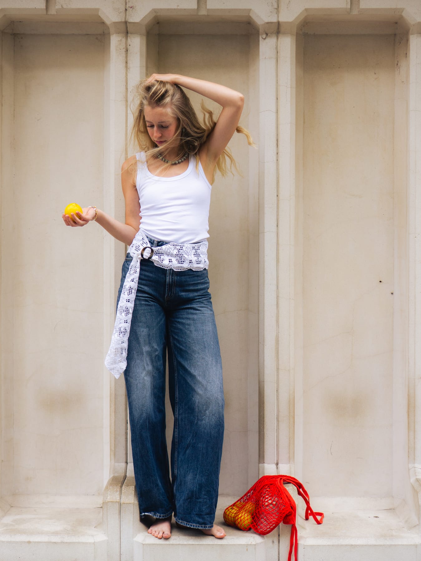 Skanzen Studio collection: model in a white top and wide jeans in a stone niche, holding a lemon, a red net bag at her feet.
