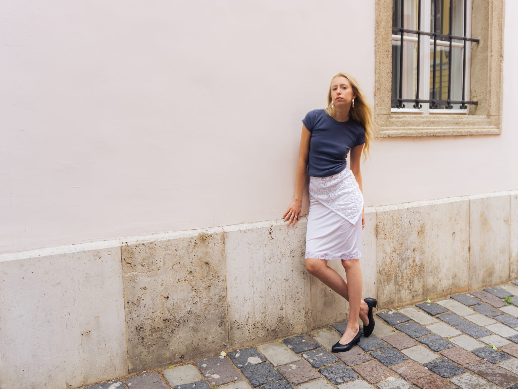 Model in a blue T-shirt and white lace slip skirt leaning on a pink wall under a barred window, Skanzen Studio.