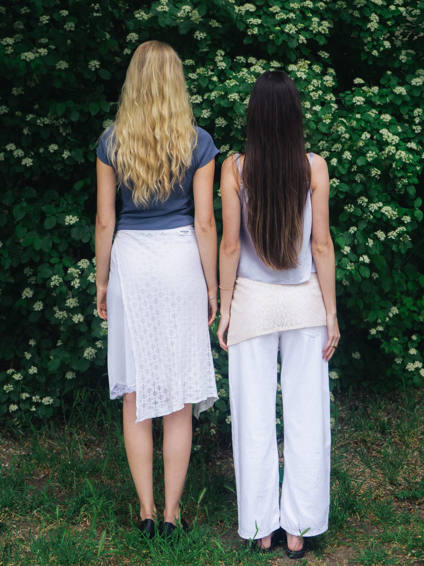 Two models seen from behind in white lace skirt and trousers in front of a flowering green hedge.