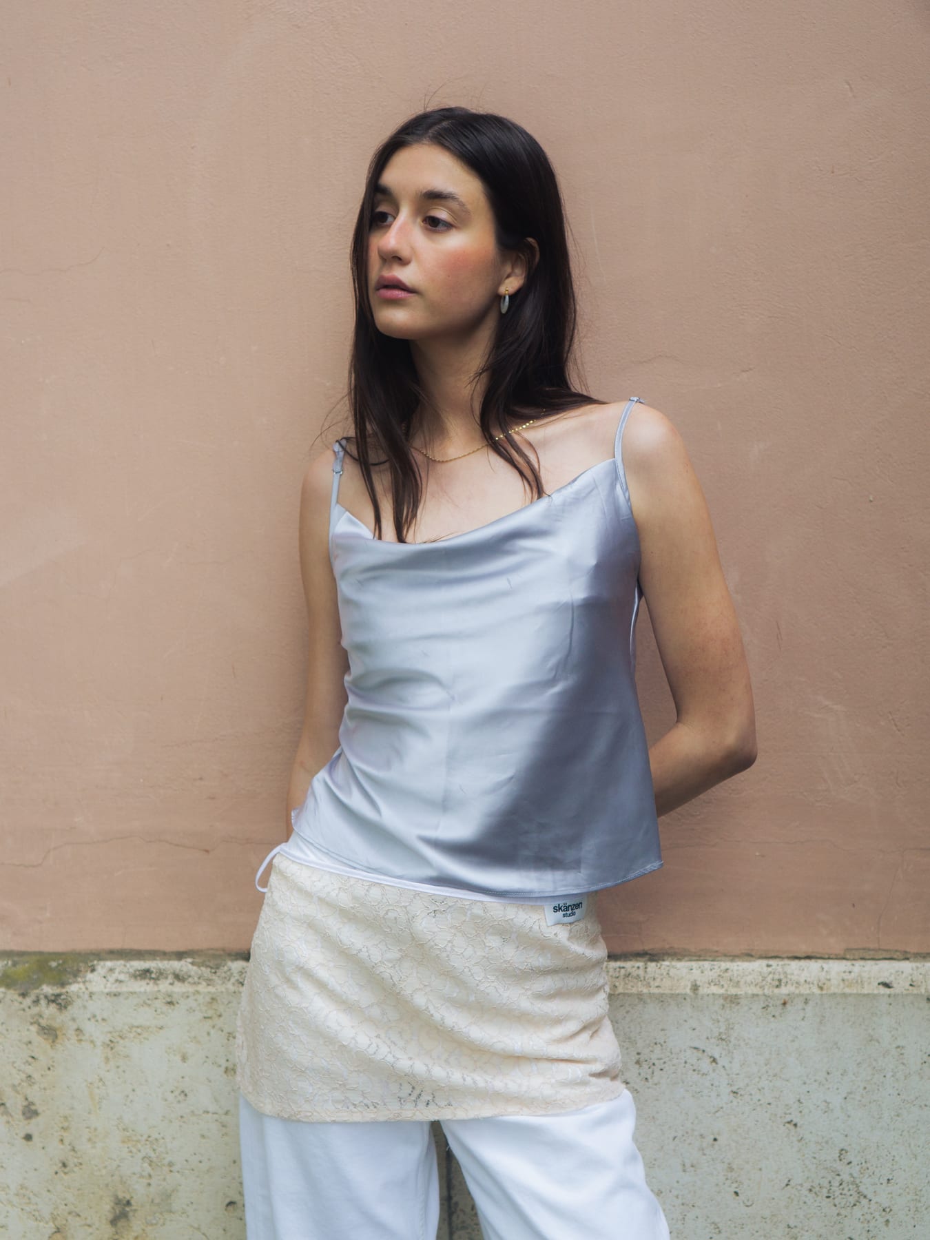 Model in a pale blue satin top and cream overskirt against a dusty pink wall.