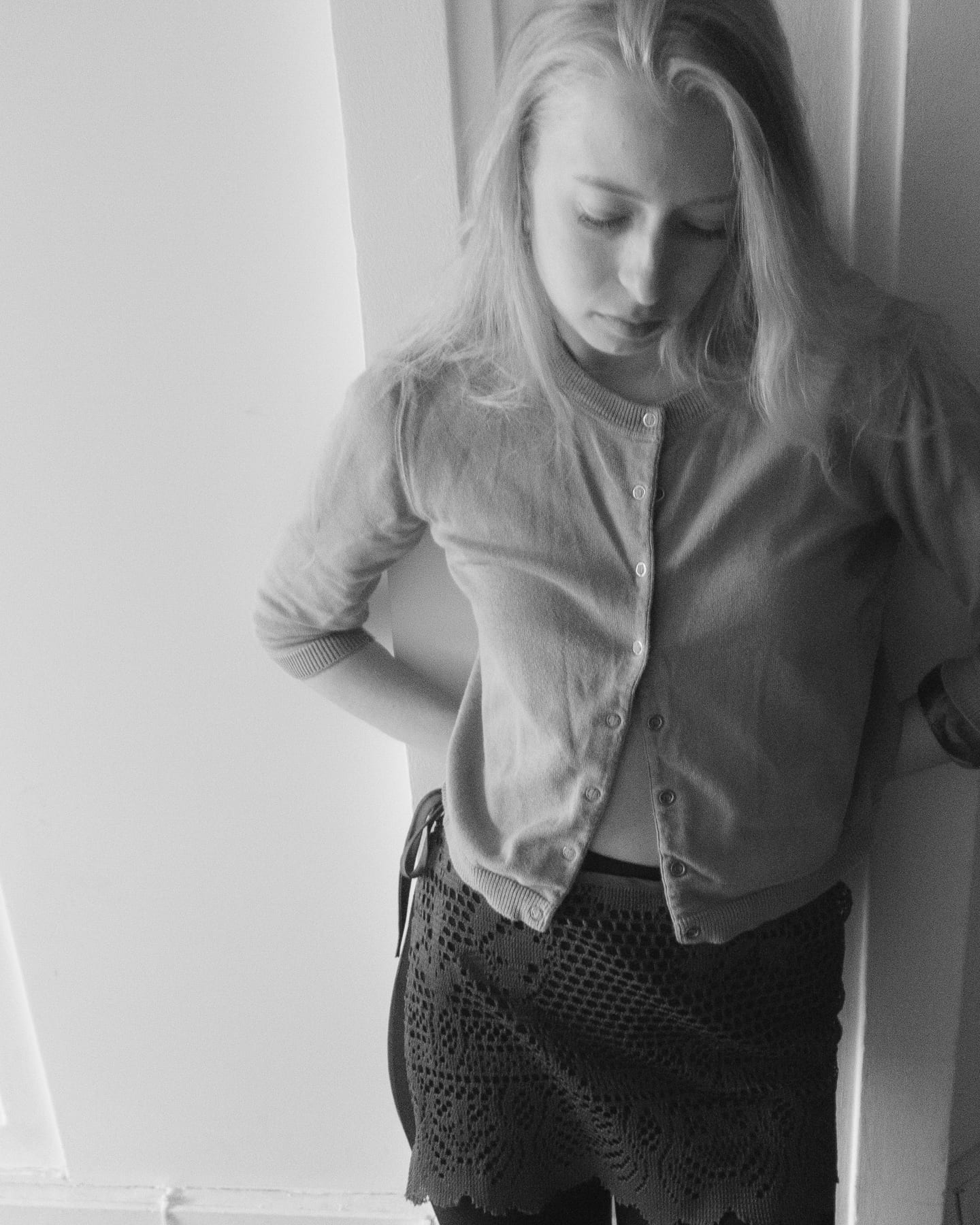 Black and white portrait of a model in a buttoned cardigan and crochet skirt leaning in a doorway, looking down.